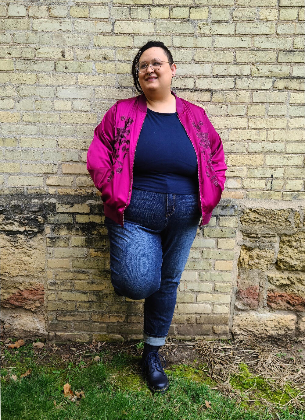 a light skinned black person in jeans and a fuchsia bomber jacket leans against a yellow brick wall and looks slightly away from camera